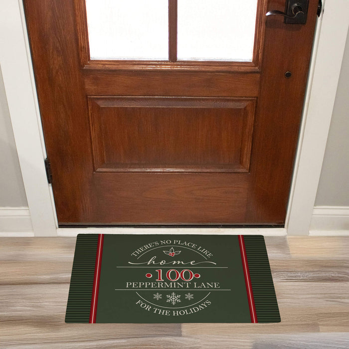 A rectangular holiday doormat placed in front of a wooden entryway door. The mat is dark green with red side stripes and white and red text reading, "There's no place like home, 100 Peppermint Lane, for the holidays," with small decorative snowflakes and holly.