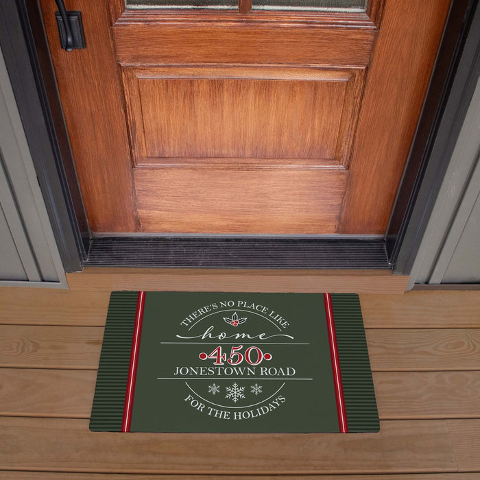 A rectangular holiday doormat placed in front of a wooden front door on a wood porch. The mat is dark green with red side stripes and white decorative text reading, "There's no place like home '450 Jonestown Road' for the holidays.