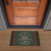 A rectangular holiday doormat placed in front of a wooden front door on a wood porch. The mat is dark green with red side stripes and white decorative text reading, "There's no place like home '450 Jonestown Road' for the holidays.