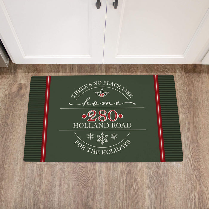 A rectangular holiday-themed doormat with a dark green background, red side stripes, and white decorative text reading 'There's no place like home ,280 Holland Road, for the holidays,' shown on a wood floor in front of white cabinet doors.
