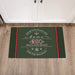 A rectangular holiday-themed doormat with a dark green background, red side stripes, and white decorative text reading 'There's no place like home ,280 Holland Road, for the holidays,' shown on a wood floor in front of white cabinet doors.