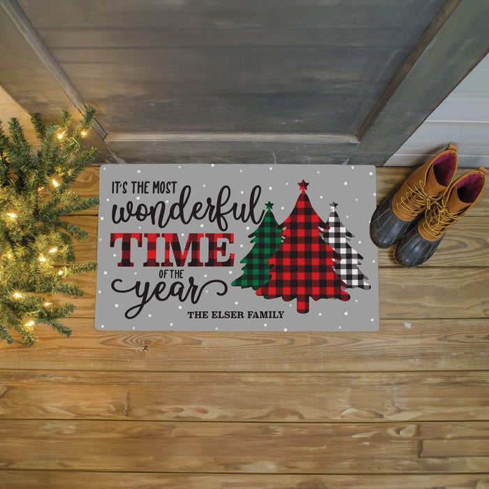 A personalized holiday doormat with the text 'IT'S THE MOST wonderful TIME OF the year' and 'THE ELSER FAMILY' above three Christmas trees in red, green, and black plaid patterns. The doormat is shown on a wooden porch floor next to a lit Christmas tree and a pair of brown boots.