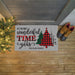 A personalized holiday doormat with the text 'IT'S THE MOST wonderful TIME OF the year' and 'THE ELSER FAMILY' above three Christmas trees in red, green, and black plaid patterns. The doormat is shown on a wooden porch floor next to a lit Christmas tree and a pair of brown boots.