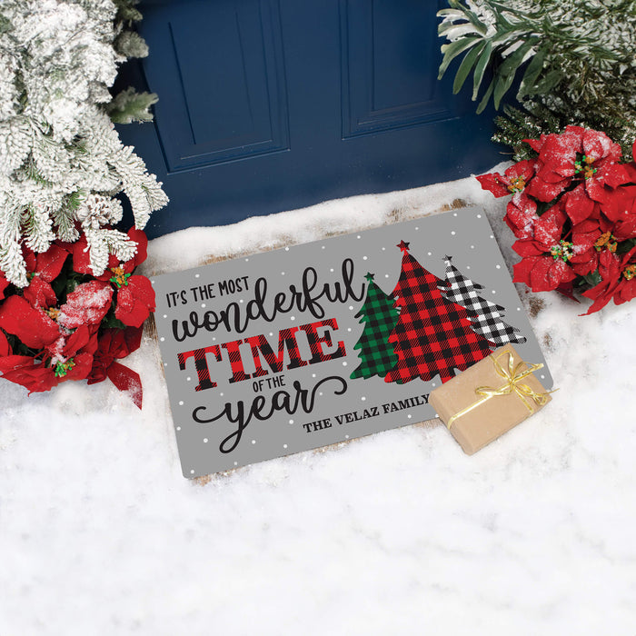 A personalized gray Christmas doormat with the text 'It's the most wonderful time of the year' and 'The Velaz Family' printed on it, styled in the snow next to a blue door with poinsettias and a small wrapped gift box.