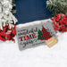A personalized gray Christmas doormat with the text 'It's the most wonderful time of the year' and 'The Velaz Family' printed on it, styled in the snow next to a blue door with poinsettias and a small wrapped gift box.