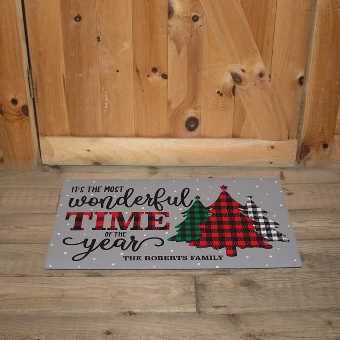 A personalized doormat with a holiday message and three Christmas tree designs in different patterns, placed on a wooden floor in front of a wooden door.
