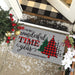 A personalized Christmas doormat with the text 'It's the most wonderful time of the year' and 'THE HARKINS FAMILY' printed on a gray background, decorated with three Christmas tree illustrations in green, red-and-black buffalo plaid, and black-and-white buffalo plaid. The mat is shown on a tiled entryway floor decorated with holiday greenery, red berries, white faux snow, and a black-and-beige buffalo plaid rug at the top.