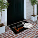 A black Halloween doormat with the words 'Welcome Boo' and a white ghost graphic, placed on a brick porch in front of a dark door with potted trees on either side.