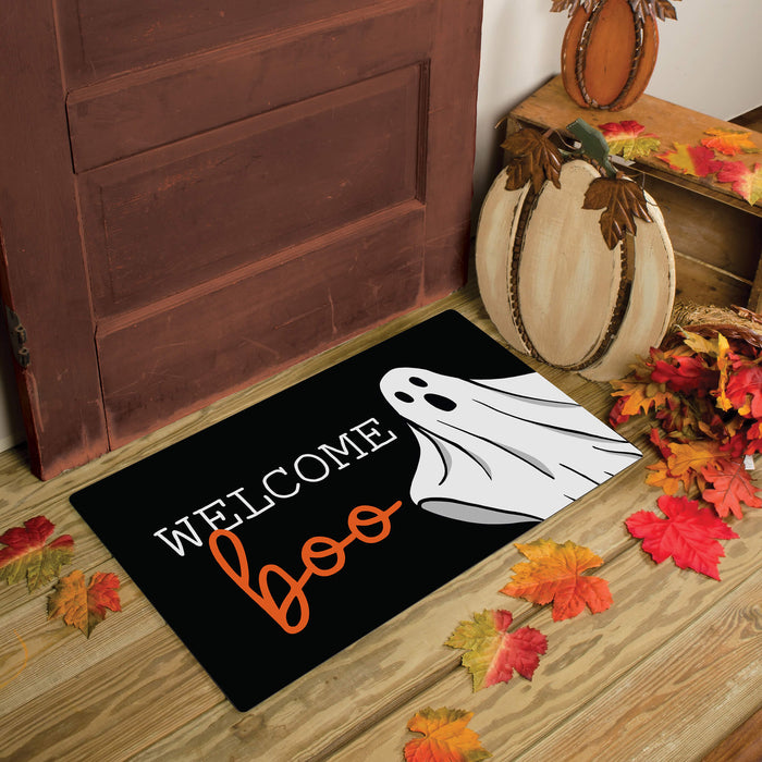 A black Halloween doormat with the words 'Welcome Boo' and a white ghost graphic, placed in front of a wooden door on a wood floor with fall leaves and pumpkins around it.