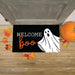A rectangular black doormat with the words “Welcome Boo” and a white ghost graphic, placed on a wooden porch floor with autumn leaves, a pumpkin, and a gray door visible.