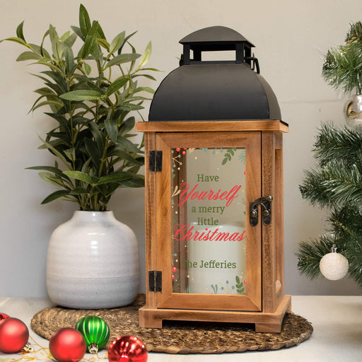 A wooden lantern with a black metal roof and glass door, decorated with a Christmas message in red and green script. The lantern is styled on a woven mat next to a potted plant and a Christmas tree branch, with red and green ornaments on the table.