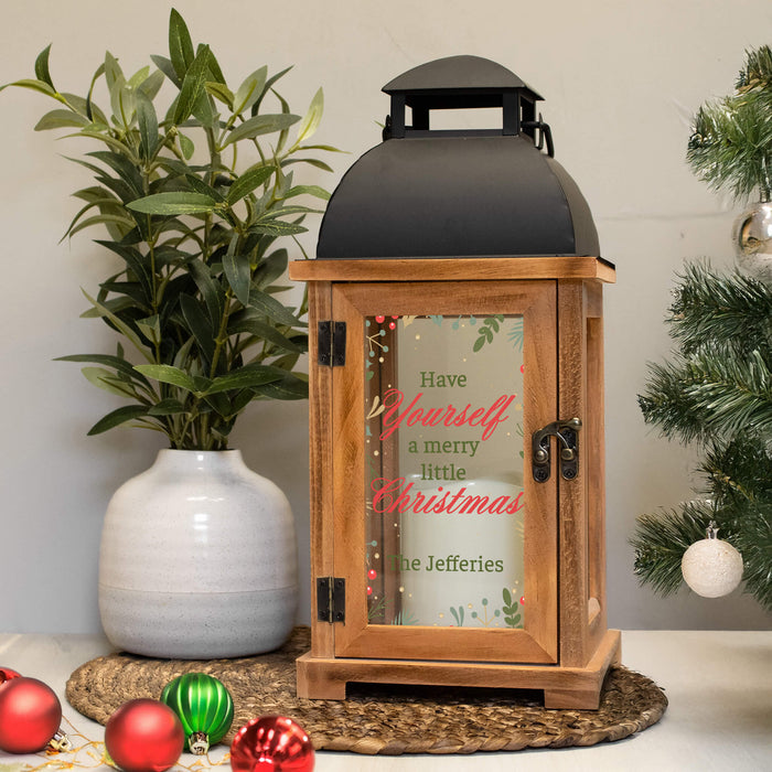 A wooden lantern with a black metal roof and glass door, decorated with a Christmas message in red and green script. The lantern is styled on a woven mat next to a potted plant and a Christmas tree branch, with red and green ornaments on the table.