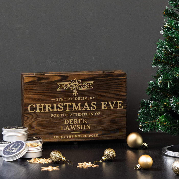 A personalized wooden Christmas Eve whiskey smoker set gift engraved with 'SPECIAL DELIVERY CHRISTMAS EVE FOR THE ATTENTION OF DEREK LAWSON FROM THE NORTH POLE' in gold lettering, styled on a dark table with gold ornaments, tins, and a Christmas tree against a dark gray background.