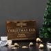 A personalized wooden Christmas Eve whiskey smoker set gift engraved with 'SPECIAL DELIVERY CHRISTMAS EVE FOR THE ATTENTION OF DEREK LAWSON FROM THE NORTH POLE' in gold lettering, styled on a dark table with gold ornaments, tins, and a Christmas tree against a dark gray background.