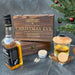 A Christmas Eve gift whiskey smoker set arranged on a dark stone surface, featuring a bottle of whiskey, a wooden engraved box labeled 'CHRISTMAS EVE' with a personalized name, a small tin of wood chips, a glass of whiskey and a wooden smoker lid, and a small wooden coaster. A decorated Christmas tree branch appears at the upper right.