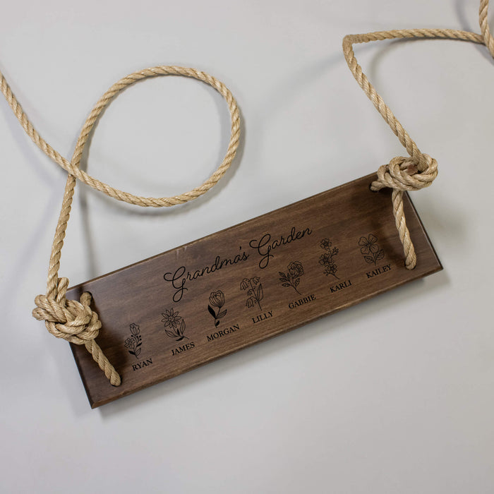Flat lay view of a wooden tree swing engraved with "Grandma's Garden" and different flower options for each grandchild's name on a gray background