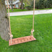 Wooden swing with engraved text hanging from a tree in a grassy area with a house across the street