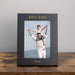 Black photo frame with a picture of a man and a boy on a wooden surface and gray background.
