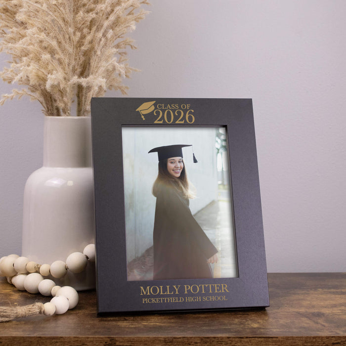 Graduation-themed photo frame with 'Class of 2026' and personalized name and school, placed on a wooden surface with decorative elements.