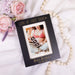 Black photo frame with a pregnancy-themed picture, flowers, and beads on a white surface