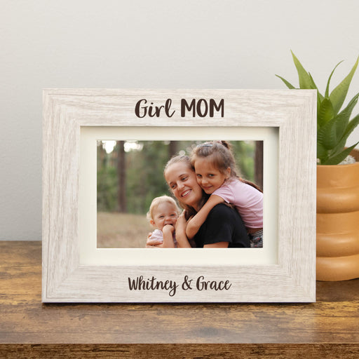 White wooden photo frame with 'Girl MOM' text, holding a family photo of a woman and two children, on a wooden surface with a plant in the background.