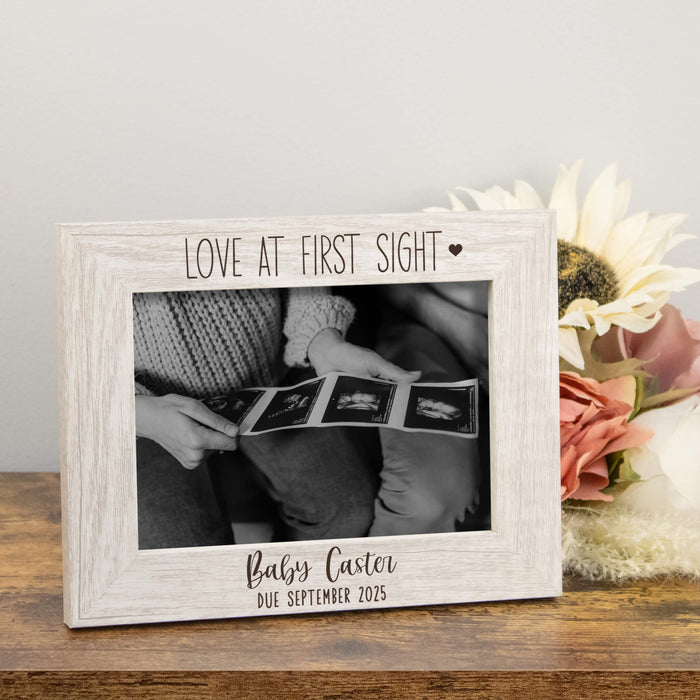 White wooden photo frame with 'Love at First Sight' text, holding ultrasound images of a baby, on a wooden surface with flowers.
