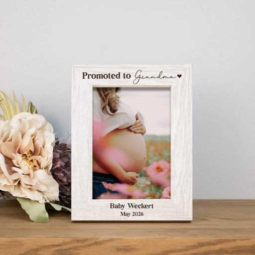 Rustic white picture frame engraved with "Promoted to Grandma" and personalization on a wooden table next to flowers