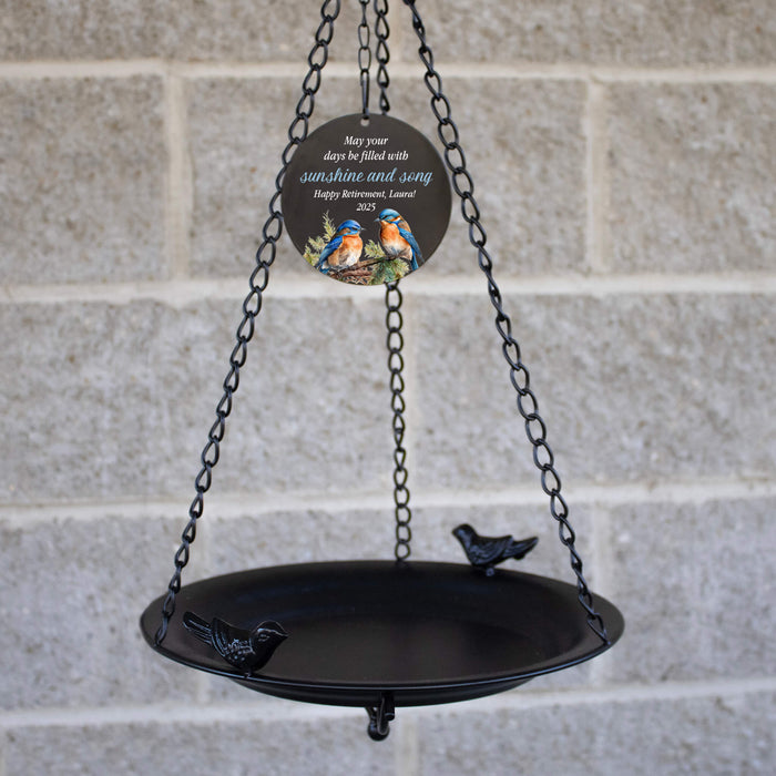 Black metal bird bath with decorative plaque against a brick wall.