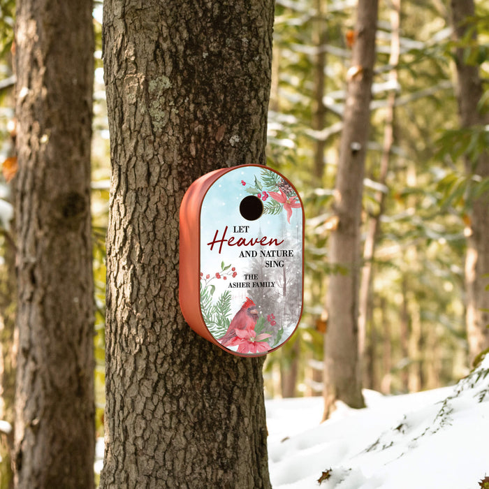 A personalized Christmas bird feeder mounted on a tree trunk in a snowy forest. The oval feeder has a red base and a printed insert featuring red cardinals, green pine branches, and the text 'Let Heaven and Nature Sing' with 'The Asher Family' at the bottom.