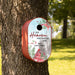 A red personalized oval birdhouse mounted on a tree trunk in a sunny backyard. The birdhouse features a red base and a printed front panel with a light blue sky, red cardinal, green leaves, and the text 'LET Heaven AND NATURE SING THE GARCIA FAMILY'.