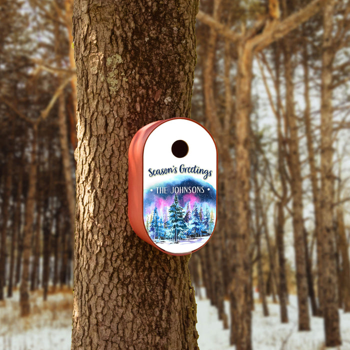 A red personalized Christmas bird house  attached to a tree trunk in a snowy forest. It features a white top section with 'Season's Greetings' and 'THE JOHNSONS' in script, and a colorful winter forest illustration below.