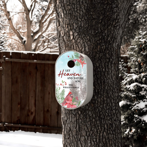 A personalized white Christmas birdhouse hanging on a tree in a snowy backyard. The birdhouse features a light blue and white background with red cardinals, green pine branches, and the text 'LET Heaven AND NATURE SING - THE WHALEN FAMILY'.