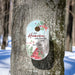 A personalized oval bird housse with a winter cardinal design, mounted on a tree trunk in a snowy outdoor setting. The house features the text 'LET Heaven AND NATURE SING' and 'THE SMITH FAMILY'.
