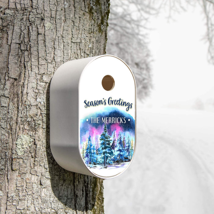 A white oval holiday bird house mounted on a tree trunk in a snowy outdoor setting. The lid features a white top section with the text 'Season's Greetings' and 'THE MERRICKS', and a colorful winter forest illustration below.