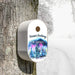 A white oval holiday bird house mounted on a tree trunk in a snowy outdoor setting. The lid features a white top section with the text 'Season's Greetings' and 'THE MERRICKS', and a colorful winter forest illustration below.