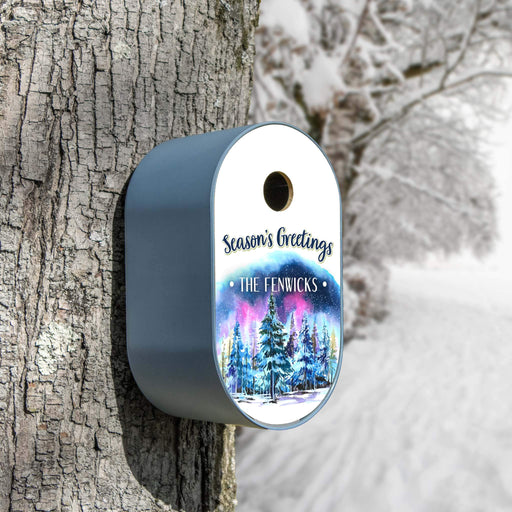 A blue personalized Christmas bird house mounted on a tree trunk in a snowy outdoor setting. The feeder has a dark gray body and a white top panel printed with 'Season's Greetings' and 'THE FENWICKS' above a colorful winter forest illustration.
