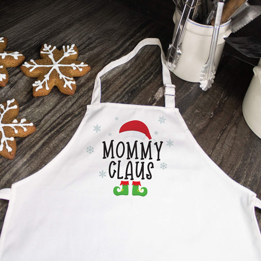A white kitchen apron with a 'Mommy Claus' holiday graphic, laid flat on a dark wooden tabletop next to gingerbread cookies and kitchen utensils.