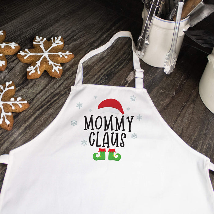 A white kitchen apron with a 'Mommy Claus' holiday graphic, laid flat on a dark wooden tabletop next to gingerbread cookies and kitchen utensils.