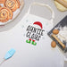 A white kitchen apron with 'AUNTIE CLAUS' printed in black, decorated with a red Santa hat, green elf boots, and light blue snowflakes. The apron is styled in a flat-lay with baking props including cinnamon rolls, eggs, flour, a brush, and dough.