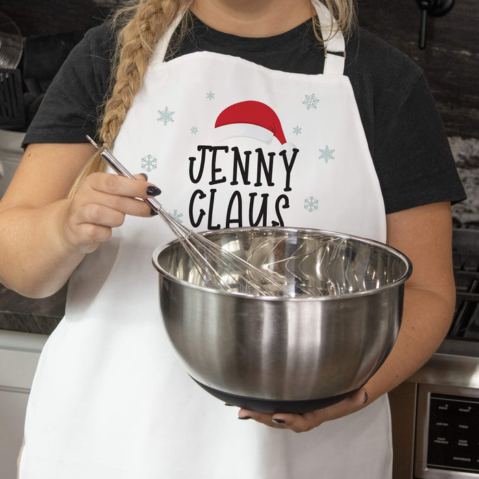 A person wearing a white apron with a red Santa hat and the text 'JENNY CLAUS' holds a stainless steel mixing bowl with a whisk in a kitchen setting.