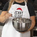 A person wearing a white apron with a red Santa hat and the text 'JENNY CLAUS' holds a stainless steel mixing bowl with a whisk in a kitchen setting.