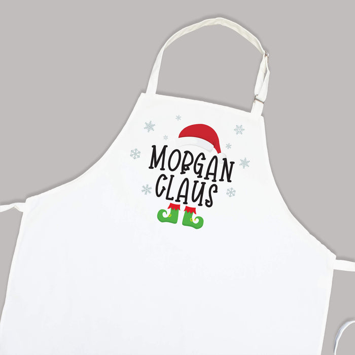 A white apron with a personalized Christmas graphic that reads 'MORGAN CLAUS' in black text, decorated with a red Santa hat, green elf boots, and light blue snowflakes, shown against a solid gray background.
