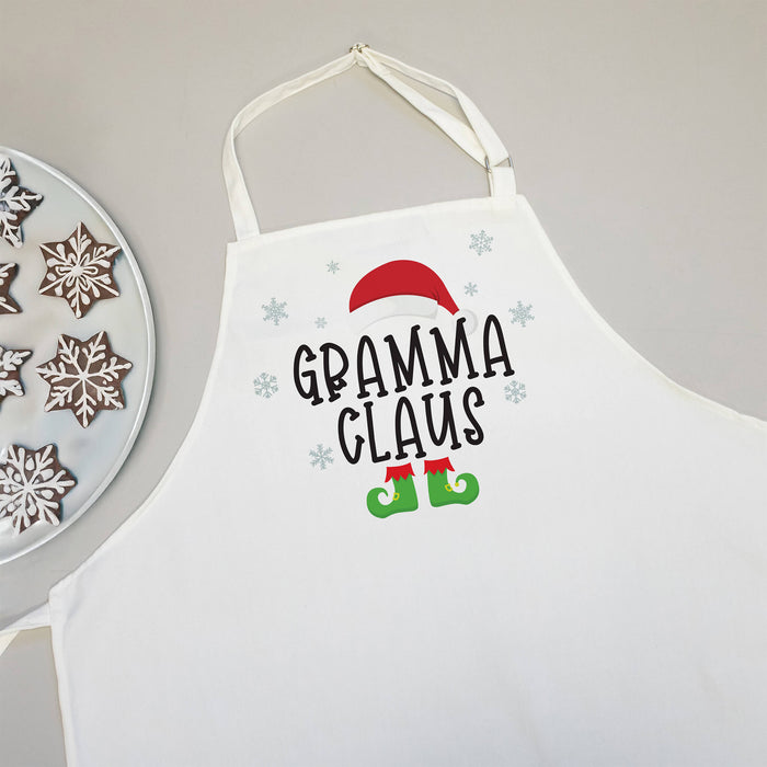 A white apron with a 'GRAMMA CLAUS' holiday graphic, featuring a red Santa hat, black text, green elf boots, and light blue snowflakes. A plate of decorated gingerbread cookies is partially visible on the left against a neutral gray background.