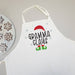 A white apron with a 'GRAMMA CLAUS' holiday graphic, featuring a red Santa hat, black text, green elf boots, and light blue snowflakes. A plate of decorated gingerbread cookies is partially visible on the left against a neutral gray background.