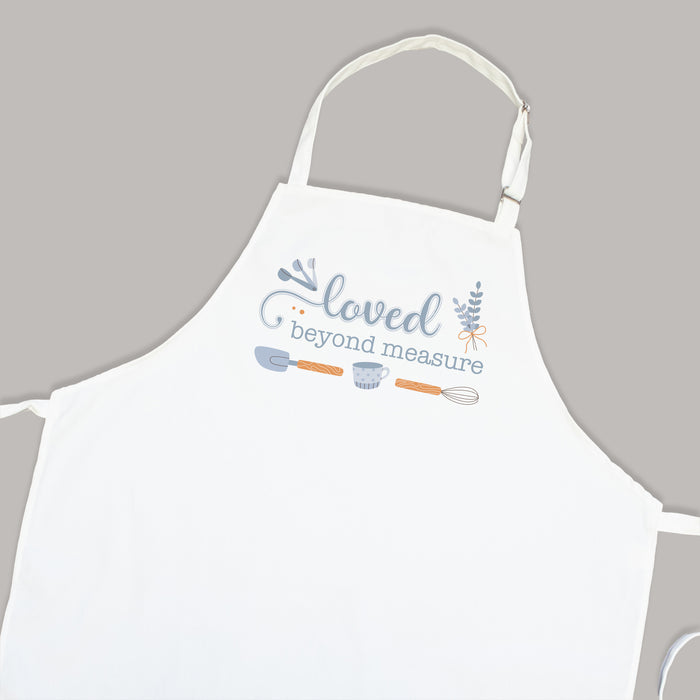 White apron on a gray background that has "loved beyond measure" and baking tools printed on it