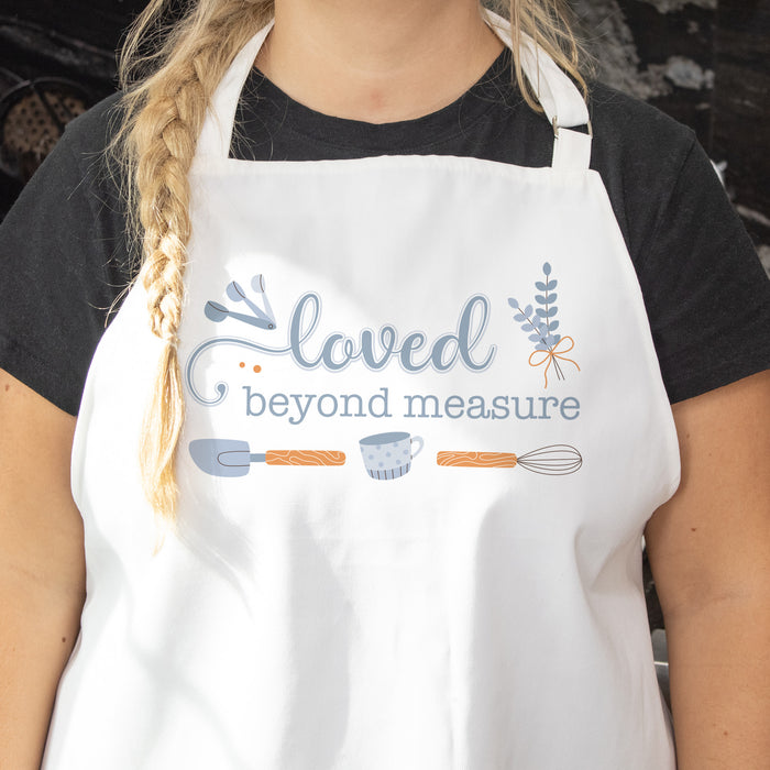 Blonde woman in a black shirt wearing a white apron that has a blue baking design printed on it