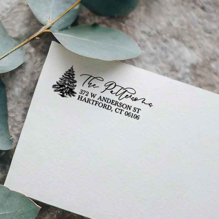 A close-up of a white textured paper envelope or card with a black printed address design featuring a pine tree and the name 'The Pettitson,' styled with eucalyptus leaves on a stone surface.