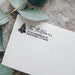 A close-up of a white textured paper envelope or card with a black printed address design featuring a pine tree and the name 'The Pettitson,' styled with eucalyptus leaves on a stone surface.