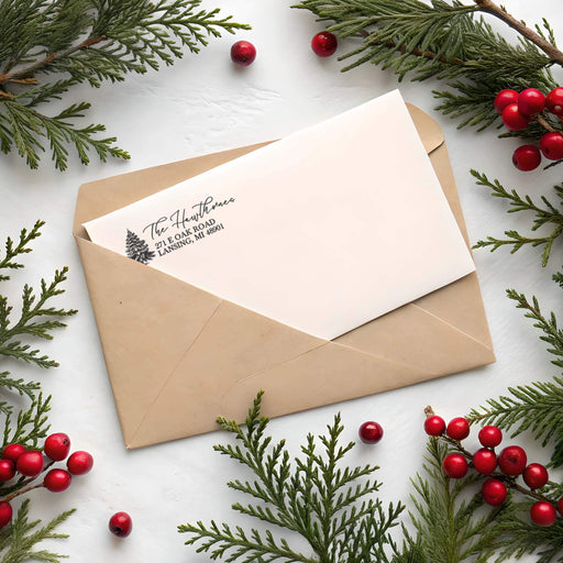 A paper envelope with a cream-colored letterhead card inserted, styled with pine branches and red berries on a white surface. The card is printed with the name 'The Hawthorne' and address text.