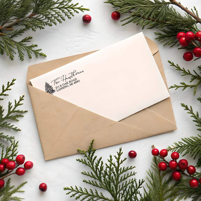 A paper envelope with a cream-colored letterhead card inserted, styled with pine branches and red berries on a white surface. The card is printed with the name 'The Hawthorne' and address text.